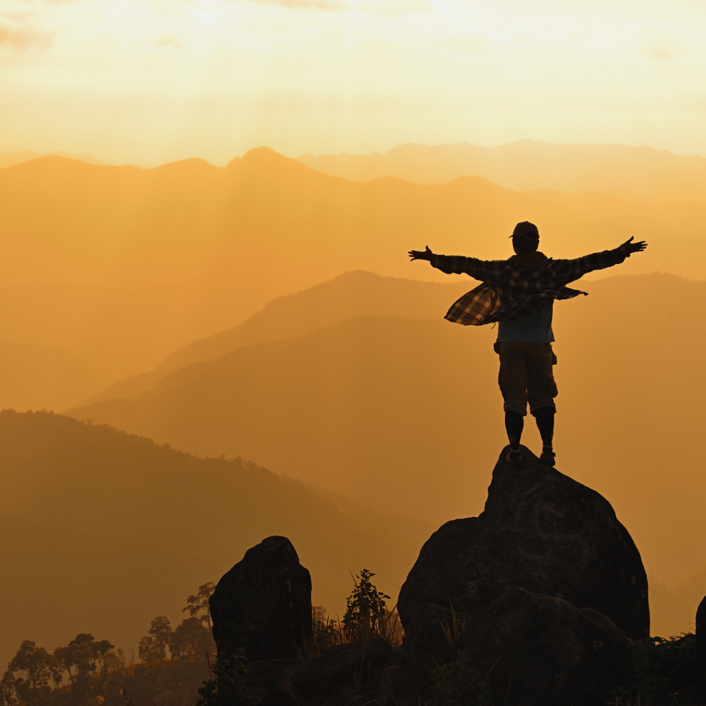 a man stands on a rock with his arms outstretched toward an orange sky