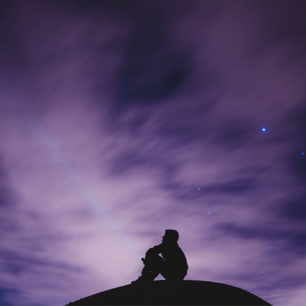 man sitting alone on a hill and behind him is a purple sky