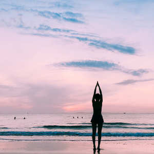 a woman performing yogic breathwork in front of the ocean and a pink sunset